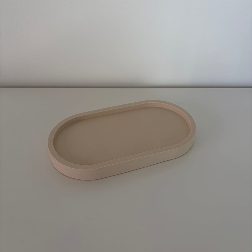 A minimalist, matte beige oval tray with raised edges, photographed on a matching light beige surface against a neutral light gray background.