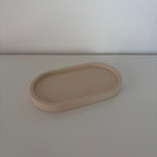 A minimalist, matte beige oval tray with raised edges, photographed on a matching light beige surface against a neutral light gray background.