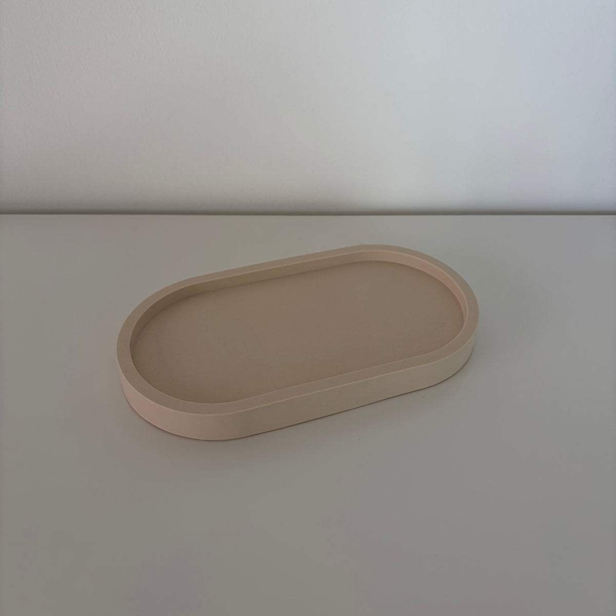 A minimalist, matte beige oval tray with raised edges, photographed on a matching light beige surface against a neutral light gray background.