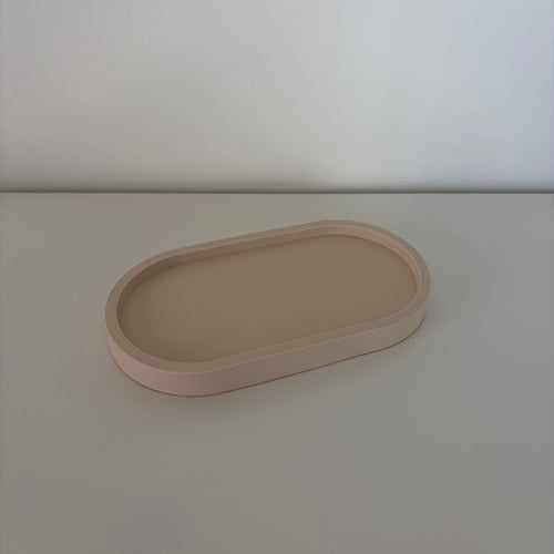 A minimalist, matte beige oval tray with raised edges, photographed on a matching light beige surface against a neutral light gray background.