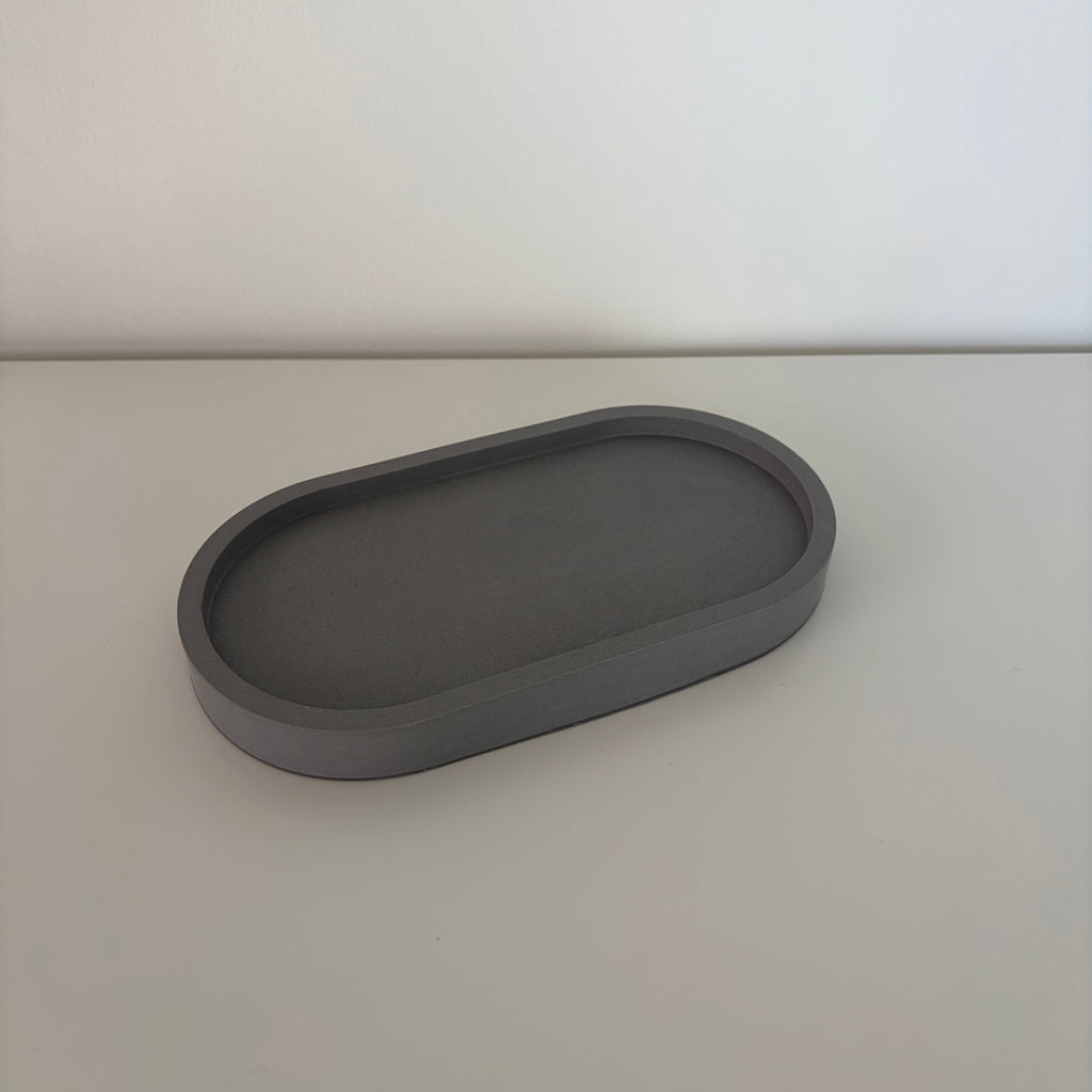 A dark gray, oval-shaped jesmonite tray with a raised rim, photographed on a light gray surface against a white background.