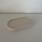 A minimalist, matte-finish oval tray in a light blush pink color, shown on a matching light gray surface against a neutral off-white background.