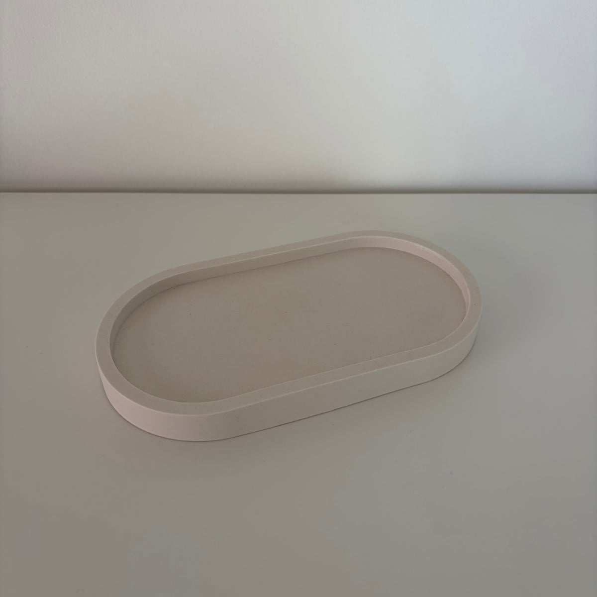 A minimalist, matte-finish oval tray in a light blush pink color, shown on a matching light gray surface against a neutral off-white background.