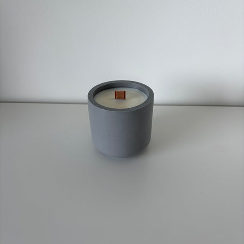 A minimalist gray ceramic candle jar with white wax and a small orange wooden wick, set against a clean, light gray background.