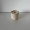 A minimalist ceramic candle jar in a warm cream-beige color with a small orange wick, set against a clean, light gray background.