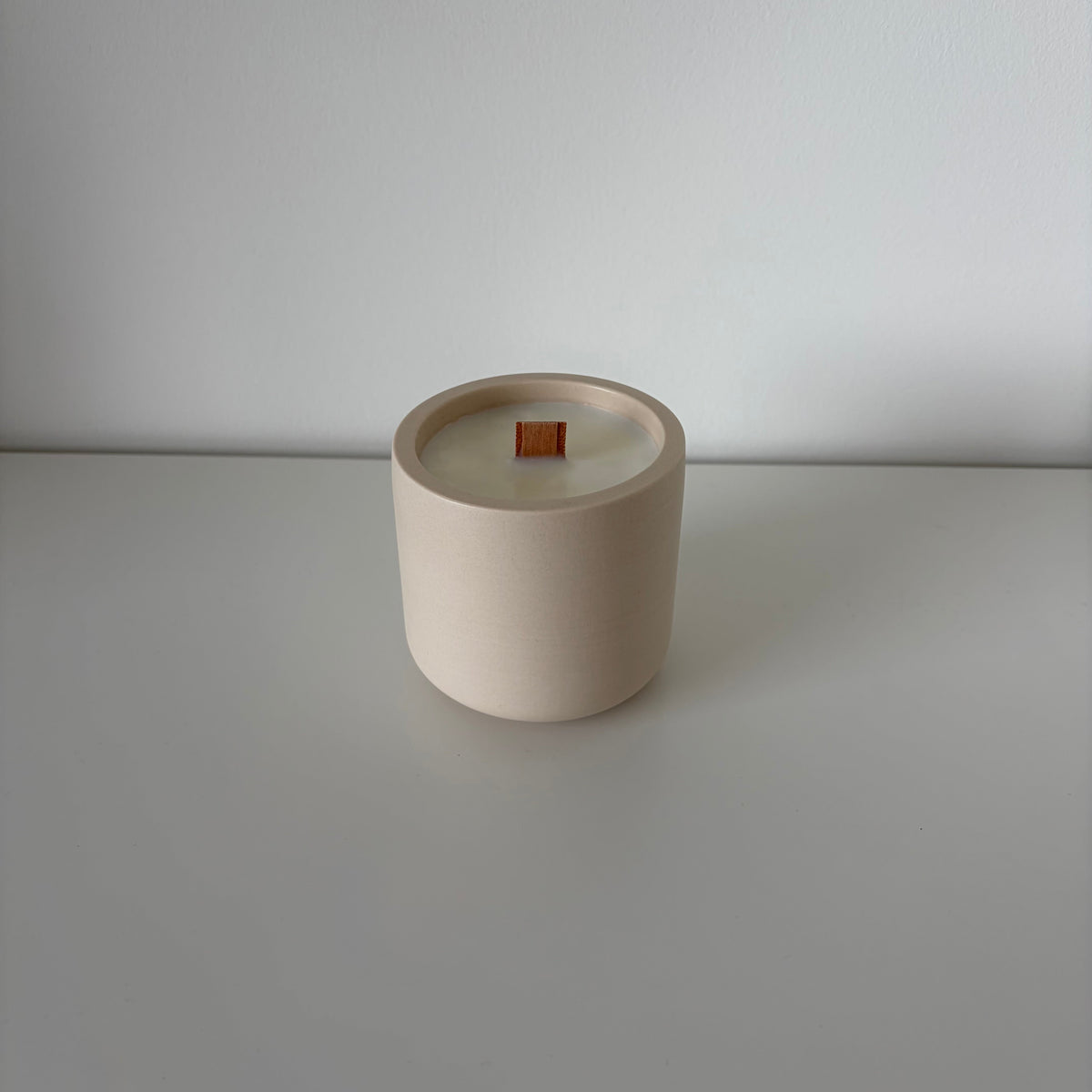 A minimalist ceramic candle jar in a warm cream-beige color with a small orange wick, set against a clean, light gray background.