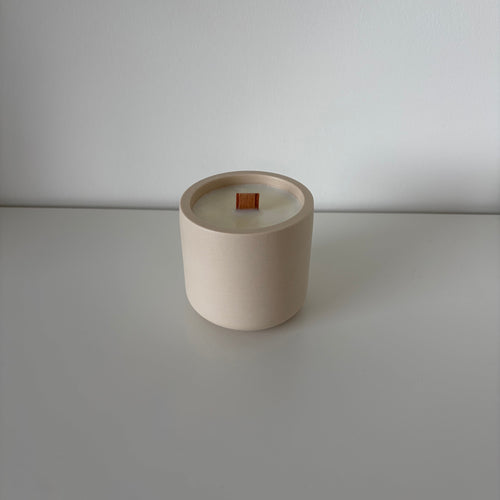 A minimalist ceramic candle jar in a warm cream-beige color with a small orange wick, set against a clean, light gray background.