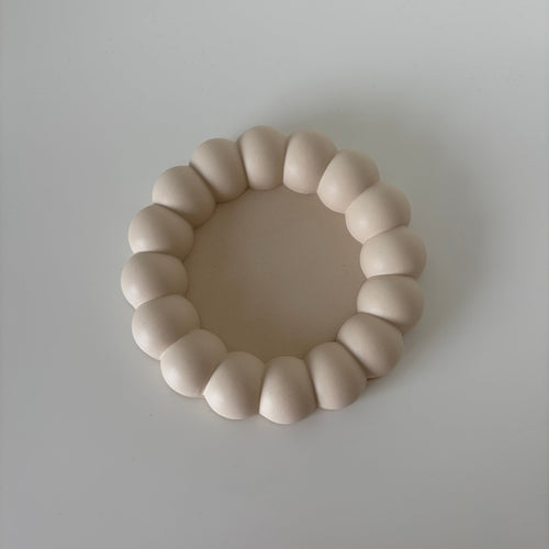 A minimalist, organic-shaped ring of smooth, rounded beige beads or tiles arranged in a circle on a light gray background.